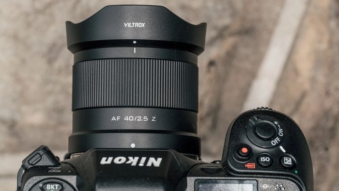 Камера Nikon Z з об'єктивом Viltrox AF 40mm f/2.5 та пелюстковою блендою лежить на бетонній поверхні вулиці, погляд зверху показує рифлене кільце фокусування під час денної документальної зйомки